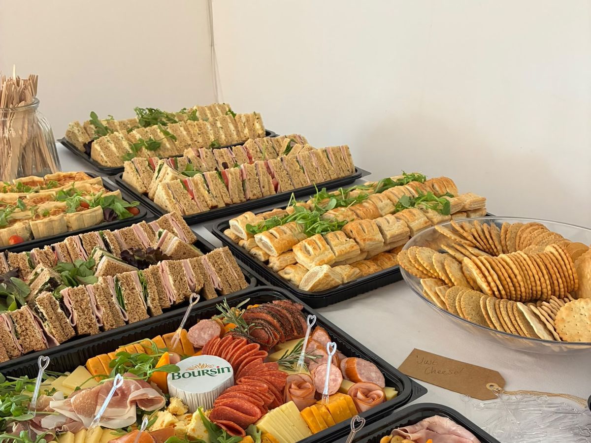 A bright buffet table with trays of freshly made sandwiches and sausage rolls beside cheese and charcuterie